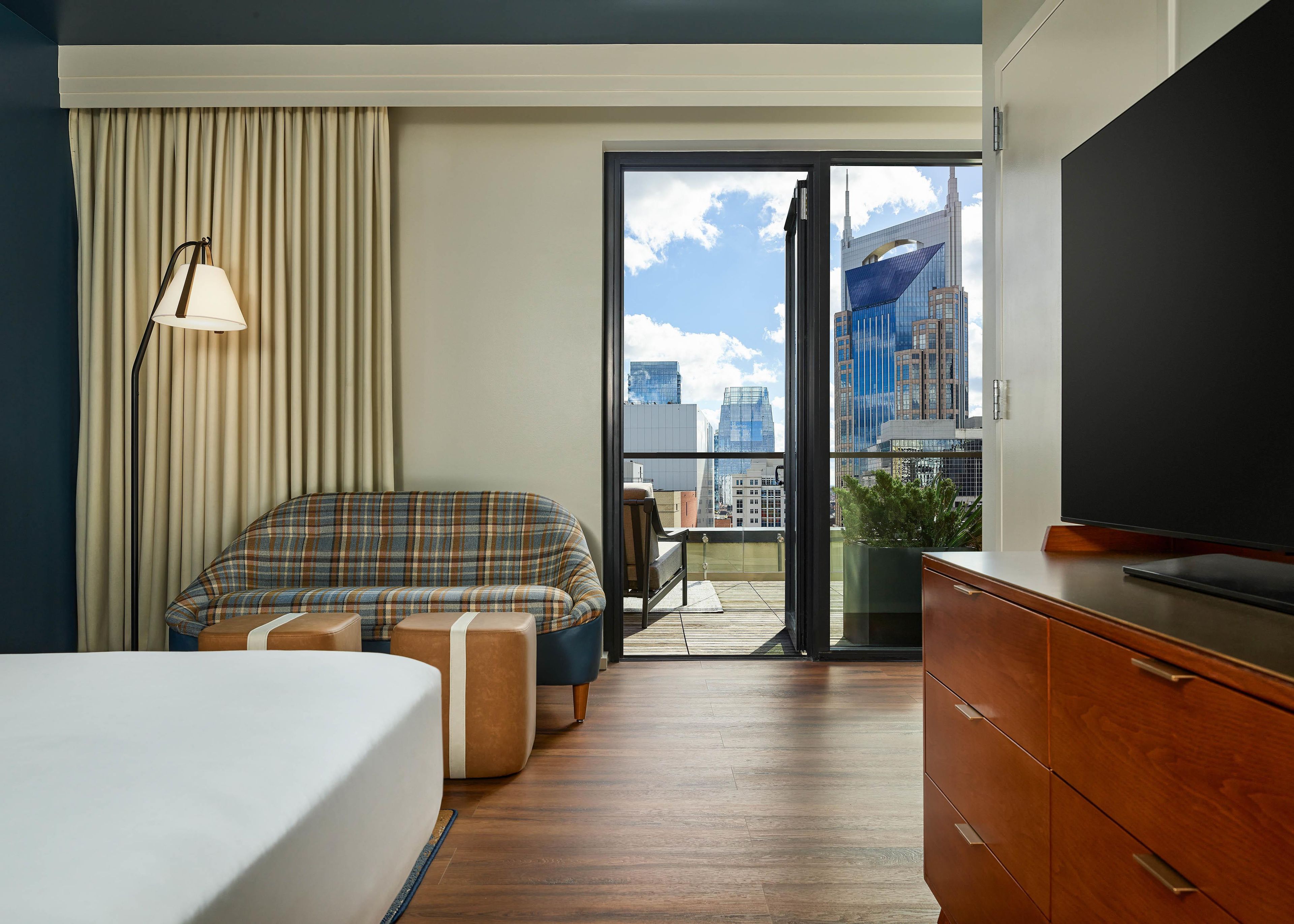 A hotel room with a bed, a sofa, a TV, a desk, a chair, a plant, and a view of the cityscape through a glass door.