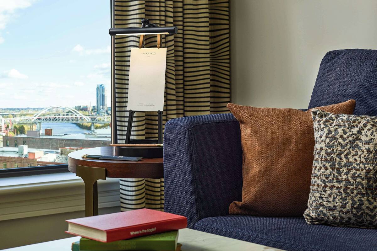 A room with a view of a bridge and river, a blue sofa with patterned cushions, and a small table with books and a sign.