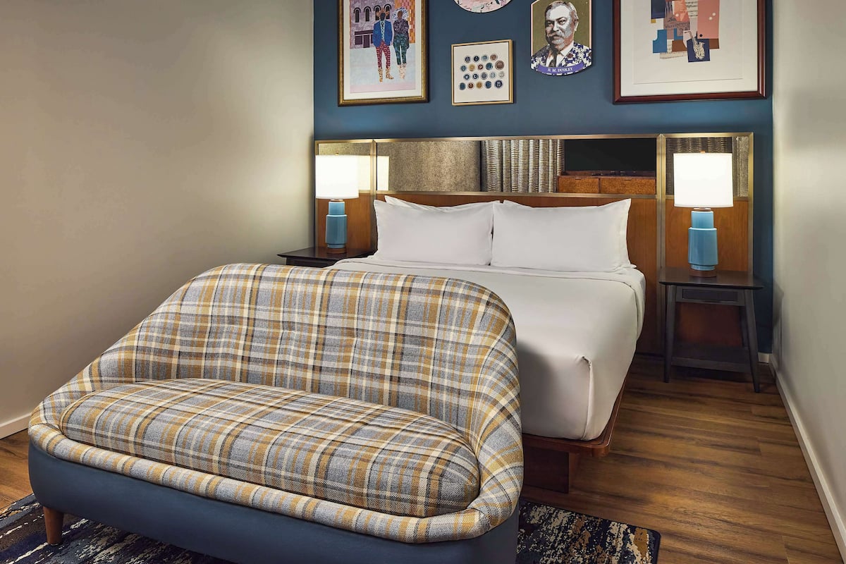A bedroom with a bed, a plaid armchair, two bedside tables with lamps, and framed artwork on the wall.