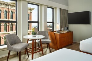 Een hotelkamer met een groot raam, een eettafel met stoelen, een houten dressoir en een bed.