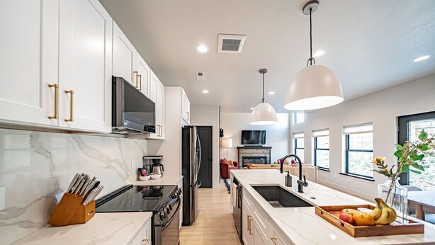 A modern kitchen with white cabinetry, a central island with a sink, and a dining area with a table and chairs. - Basecamp Stays: Stylish Home - Hot Tub, AC, Garage (Driggs)