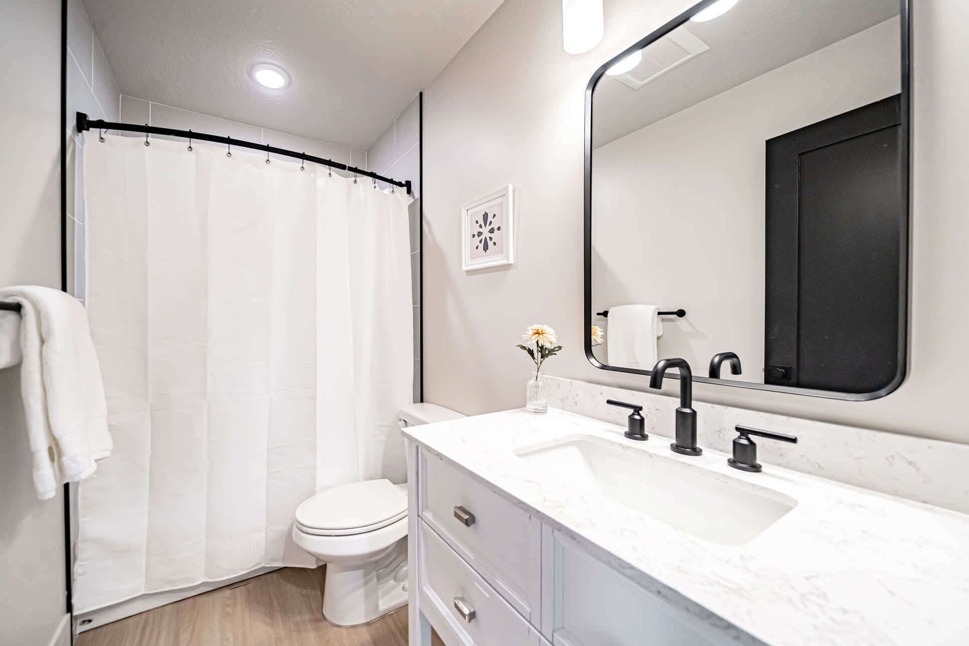 Une salle de bain moderne avec un rideau de douche, des toilettes, un lavabo doté de robinets noirs et un grand miroir.