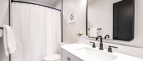 Une salle de bain moderne avec un rideau de douche, des toilettes, un lavabo doté de robinets noirs et un grand miroir.