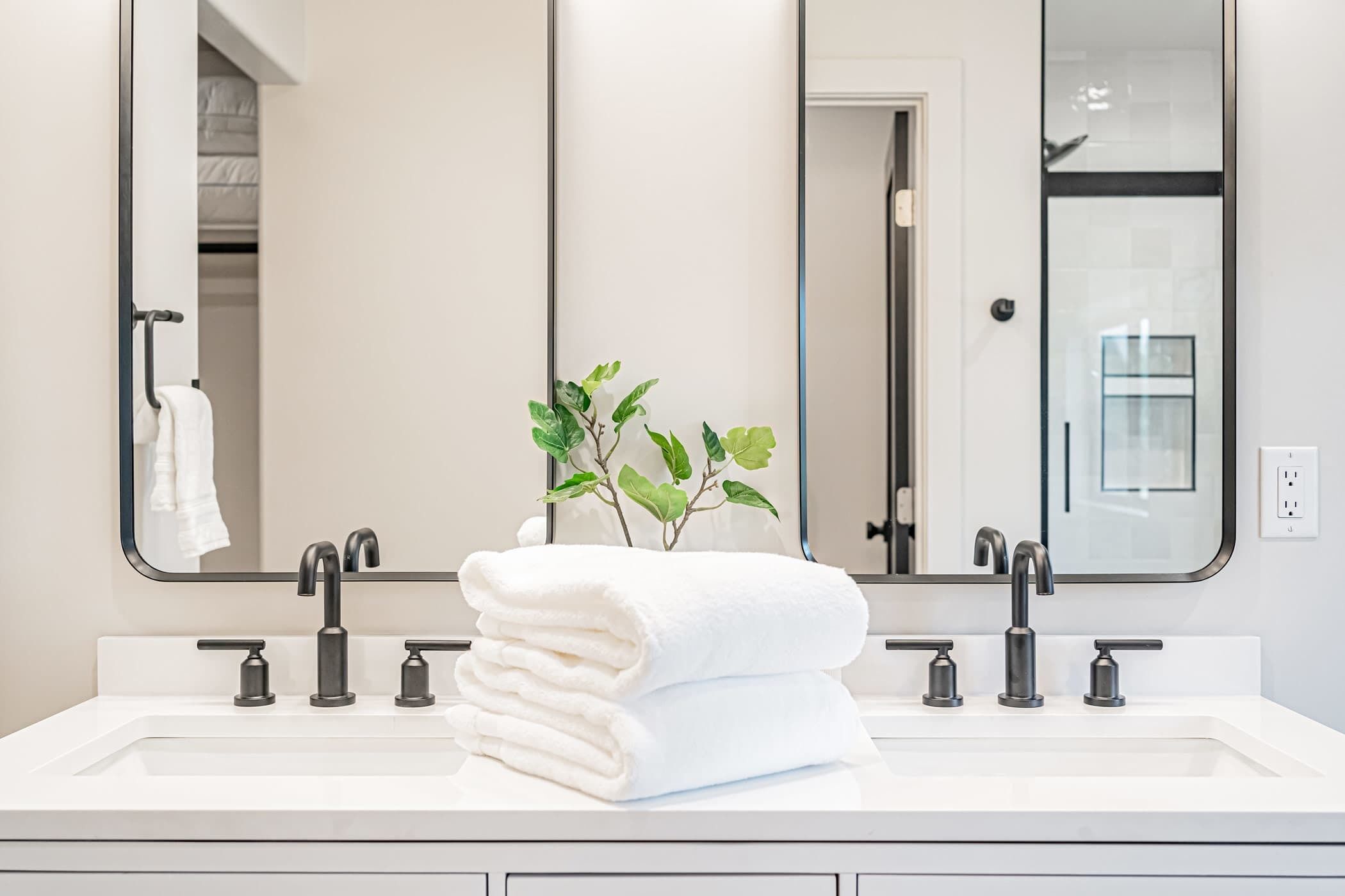 A modern bathroom with a white sink, two faucets, and a mirror. There is a plant and a stack of white towels on the sink.