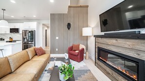 A modern living room with a sofa, a coffee table with a plant, a red armchair, a fireplace, and a TV mounted on the wall. - Basecamp Stays: Stylish Home - Hot Tub, AC, Garage (Driggs)