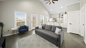 A spacious living room with a gray sofa, a blue armchair, and a patterned rug. There is a ceiling fan, a white door labeled "BEACH HOUSE," and a view of the outside through a window with blinds. - Brand New Home! Beach Views! Sleeps 6! Pets Welcome! (Bolivar Peninsula)