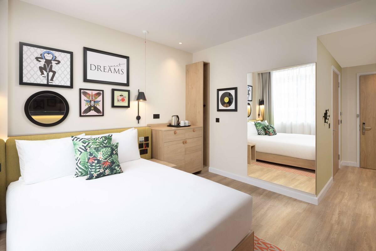 A modern hotel room with a large bed, a nightstand, a wooden wardrobe, and a mirror on the wall.