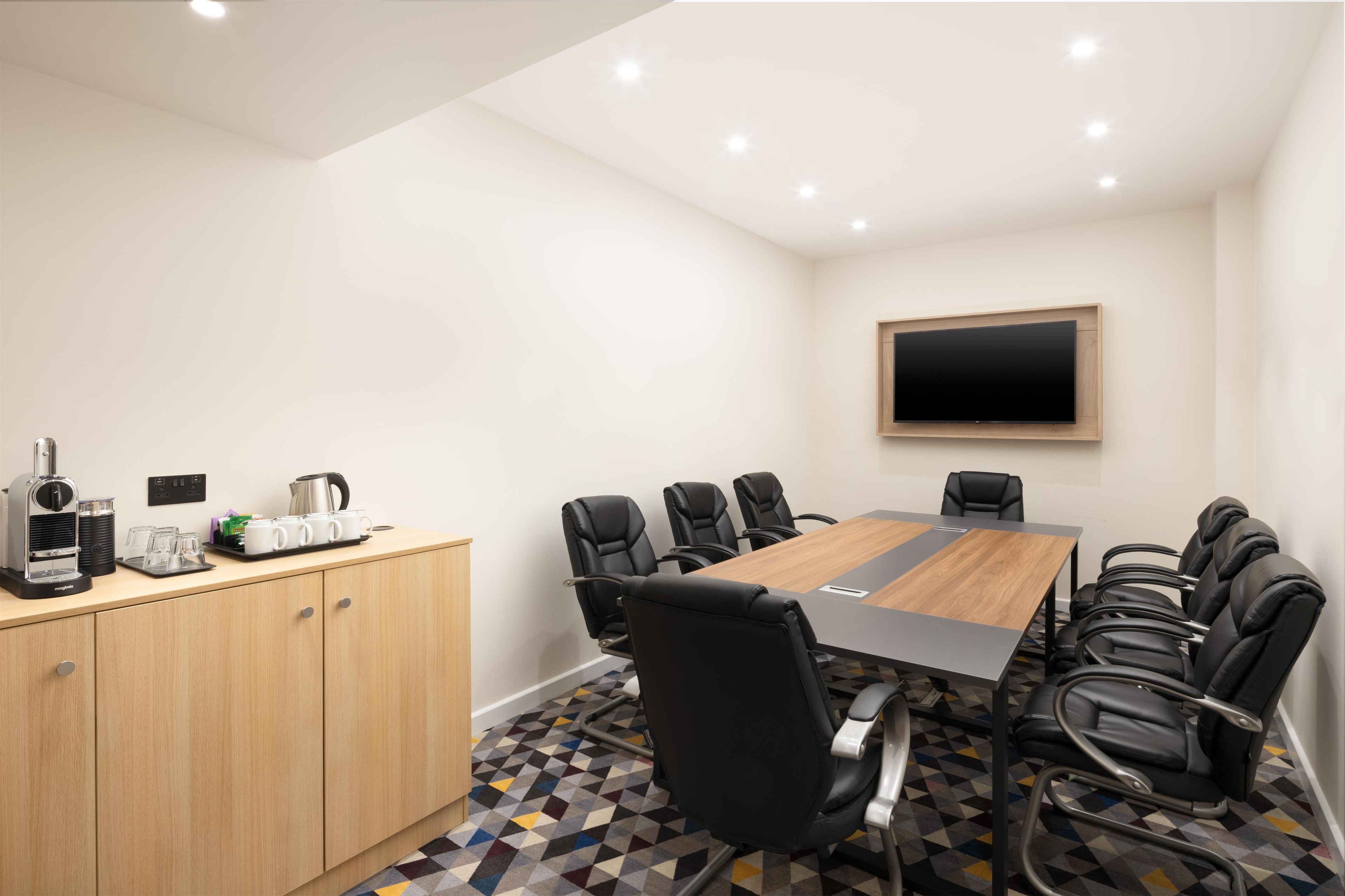 Uma sala de conferências com uma mesa comprida, cadeiras pretas, uma máquina de café e uma TV na parede.