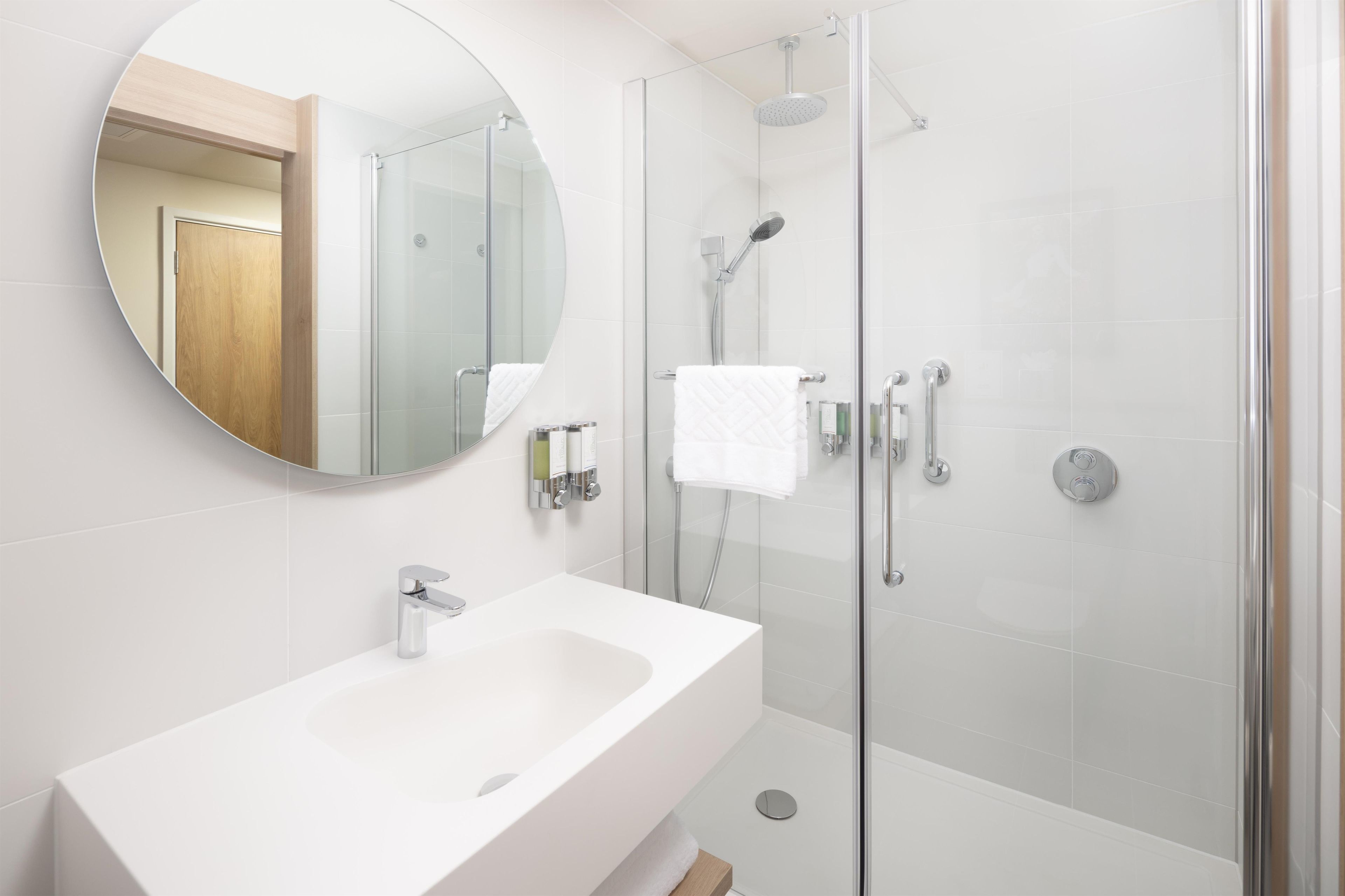 Un baño moderno con ducha envuelta en vidrio, un lavamanos blanco y un espejo redondo.