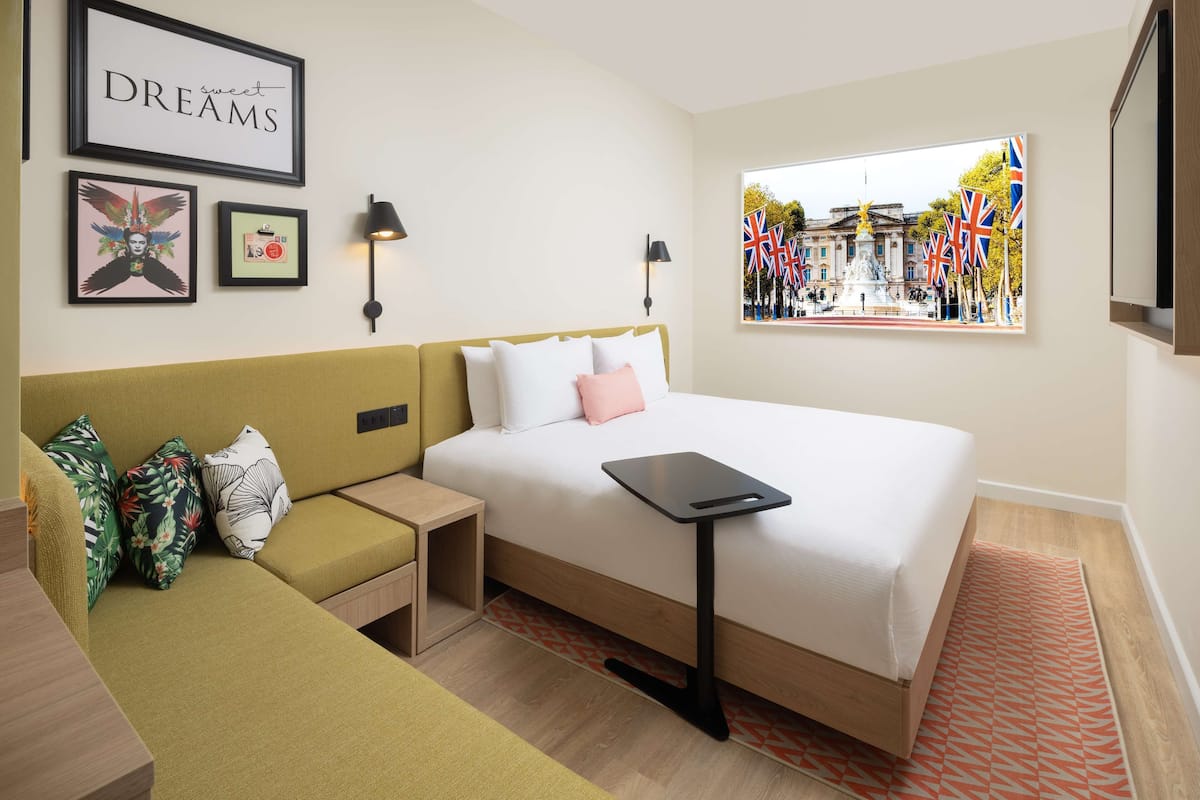 A hotel room with a bed, a nightstand, a sofa, and a wall with framed pictures and a window view of a cityscape.