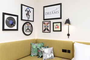 A living room with a mustard-colored sofa, a black floor lamp, and various framed artworks on the wall. - Hampton By Hilton London Old Street (London)