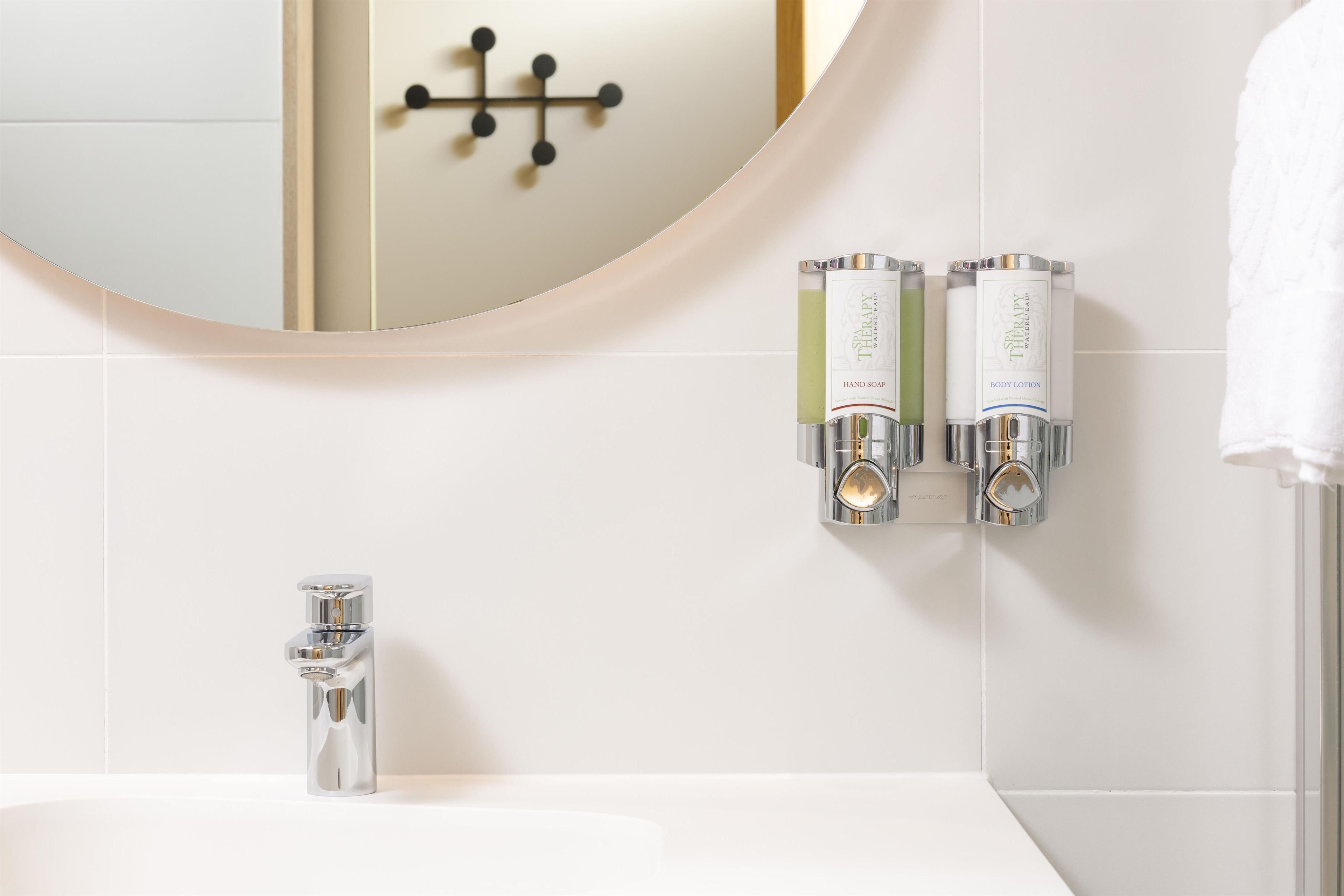 A bathroom with a round mirror, a chrome faucet, and two wall-mounted dispensers for hand soap and body lotion.