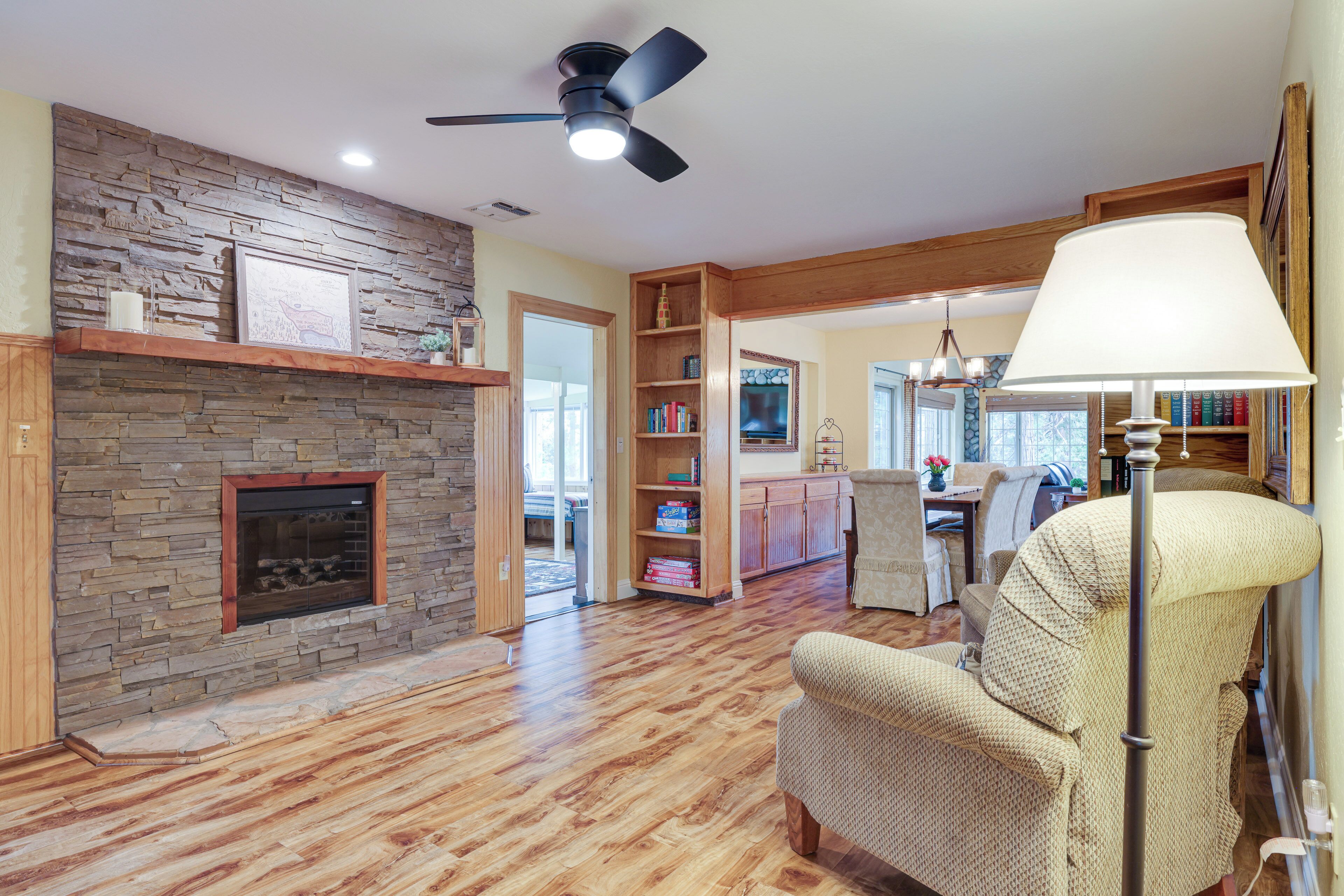 A spacious living room with a stone fireplace, wooden flooring, a ceiling fan, and a large window.