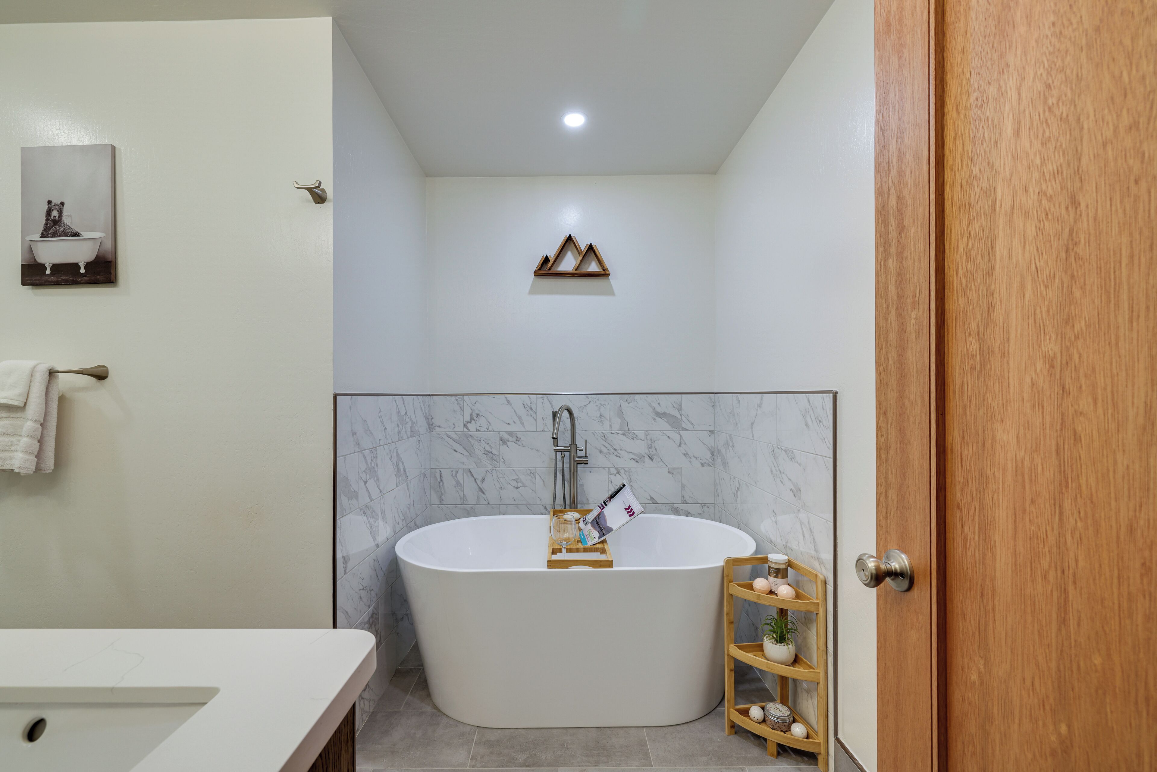 A modern bathroom with a freestanding bathtub, a wooden shelf with toiletries, and a marble wall.