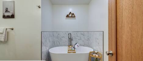A modern bathroom with a freestanding bathtub, a wooden shelf with toiletries, and a marble wall.