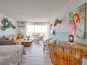 A spacious living room with a large window, a sofa, a dining table, and a wicker chair.