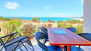 Mesa de madera con sillas azules y un cojín negro, con vista a una playa de aguas azules y tierra distante.