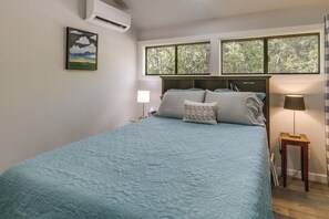 A bedroom with a bed, two bedside lamps, a window with a view of trees, and an air conditioning unit on the wall.