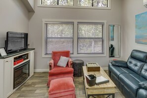 Una sala de estar con chimenea, un sillón rojo, un sofá de cuero negro y un ventanal con persianas.