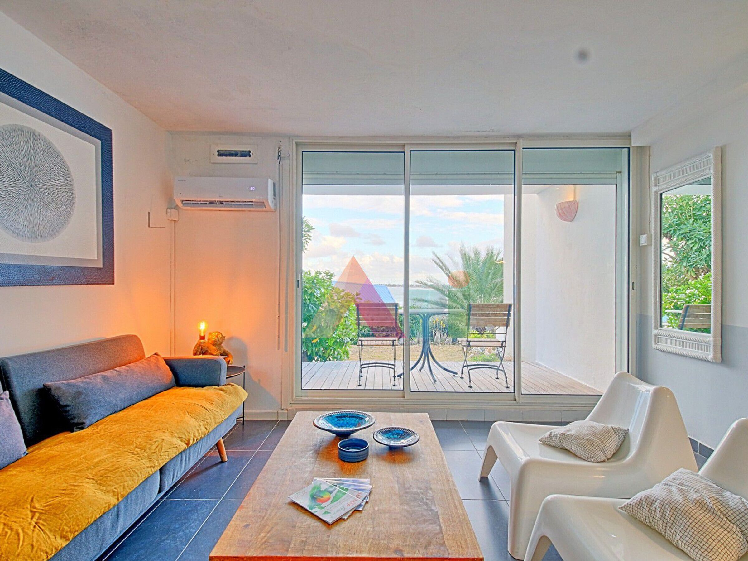 A modern living room with a sofa, coffee table, and chairs, large sliding glass doors to a balcony with outdoor furniture.