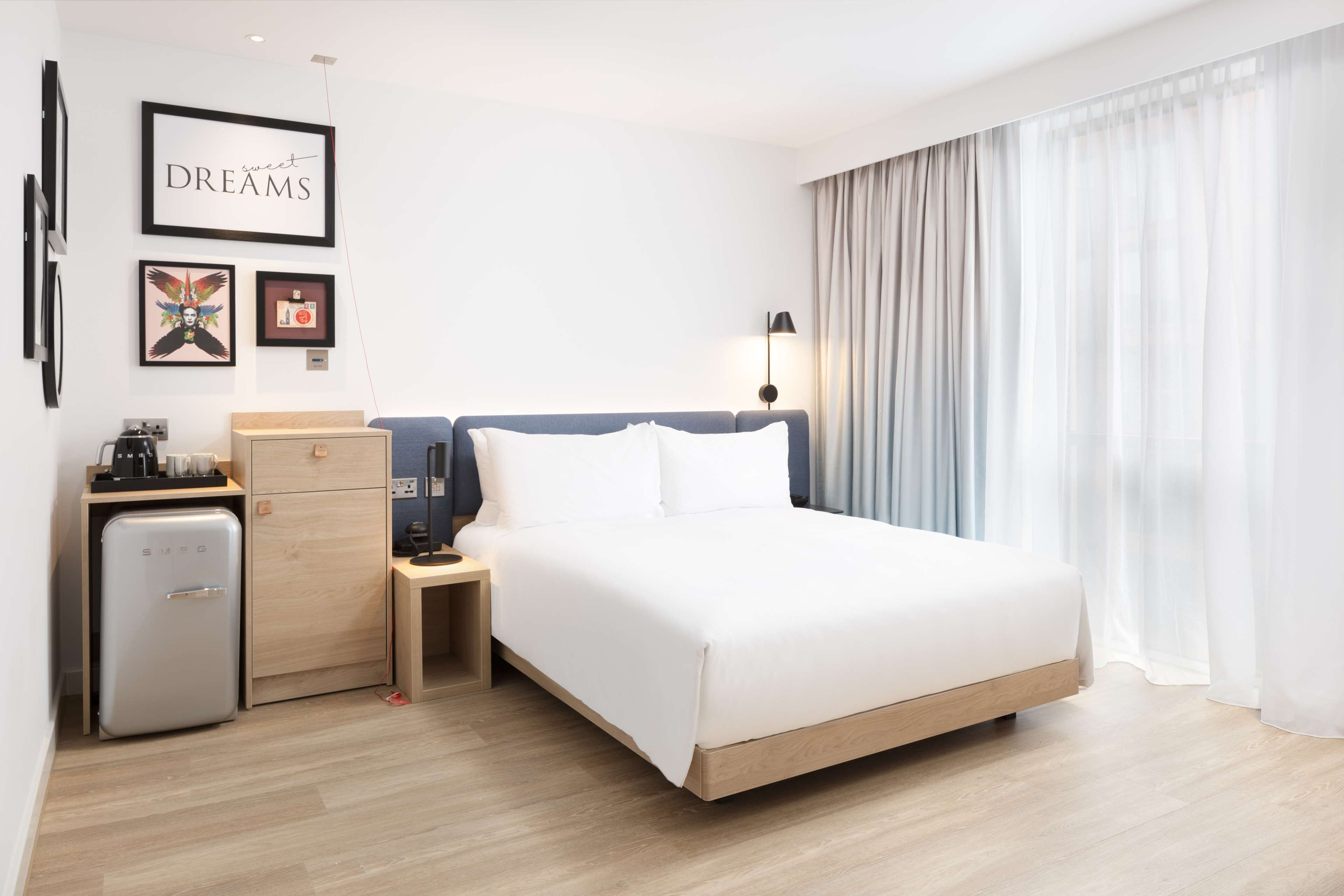 A modern hotel room with a large bed, a desk with a lamp, a minibar, and a view of the city through sheer curtains.