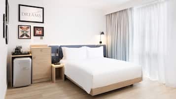 A modern hotel room with a large bed, a desk with a lamp, a minibar, and a view of the city through sheer curtains.