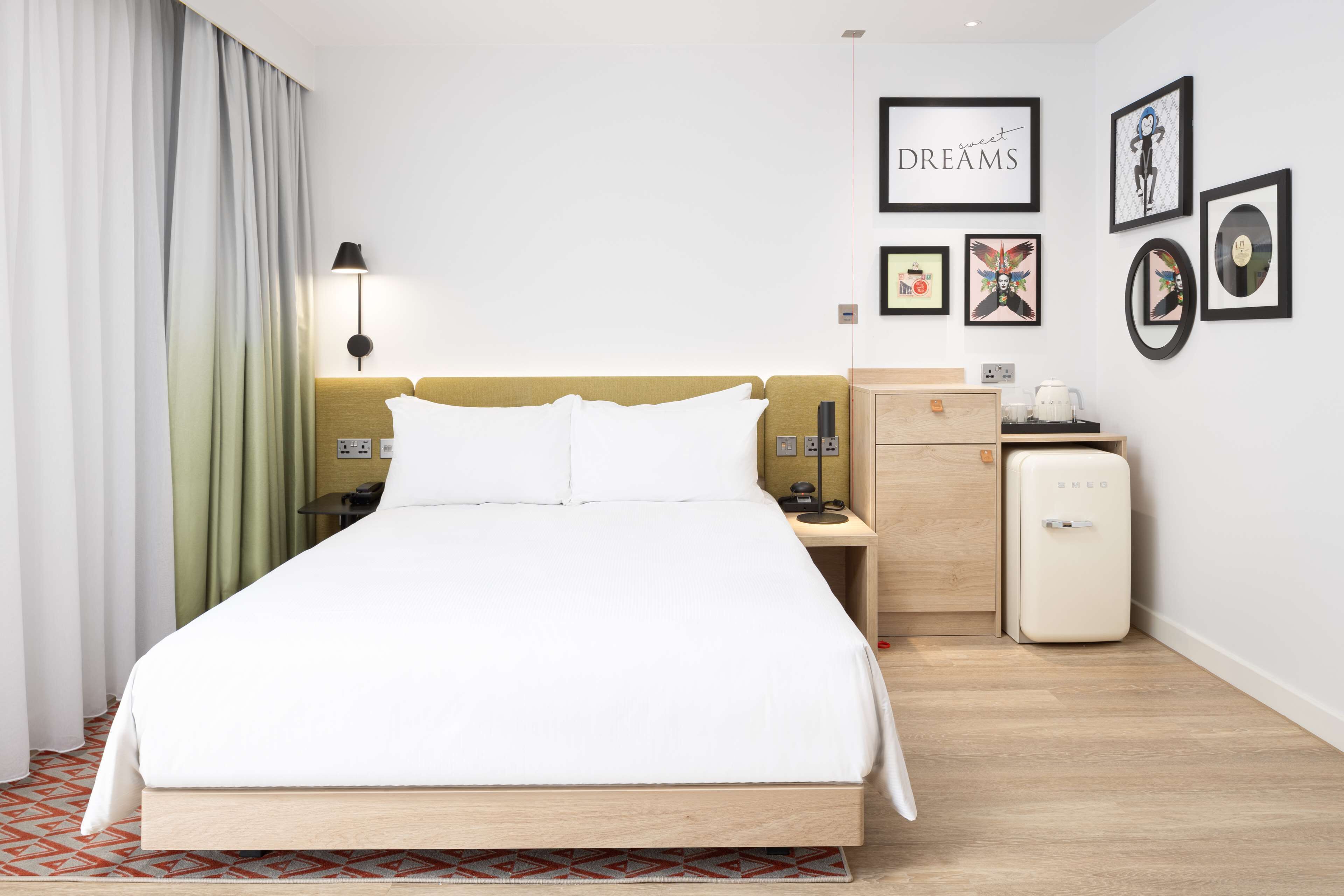 A hotel room with a bed, a nightstand, a lamp, a dresser, and a refrigerator.