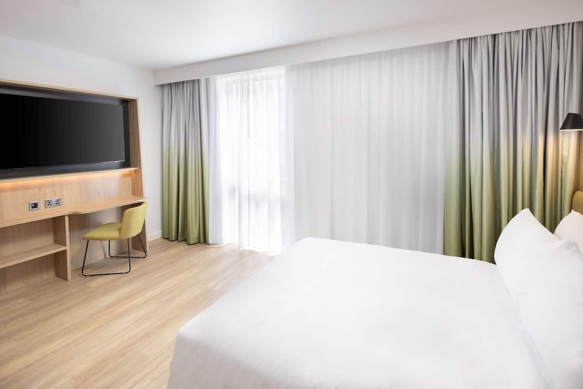 A hotel room with a bed, a desk with a chair, a television, and large windows with curtains.