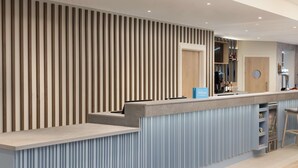 A modern hotel reception area with a wooden slat wall, a counter with a blue sign, and a bar area with a minibar. - Hampton By Hilton Rochdale (Rochdale)