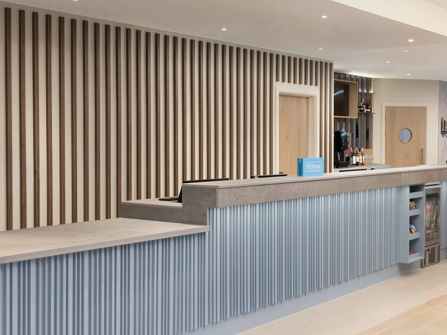 Een moderne hotel receptie met een wand van houten latten, een balie met een blauw bord en een bar met een minibar.