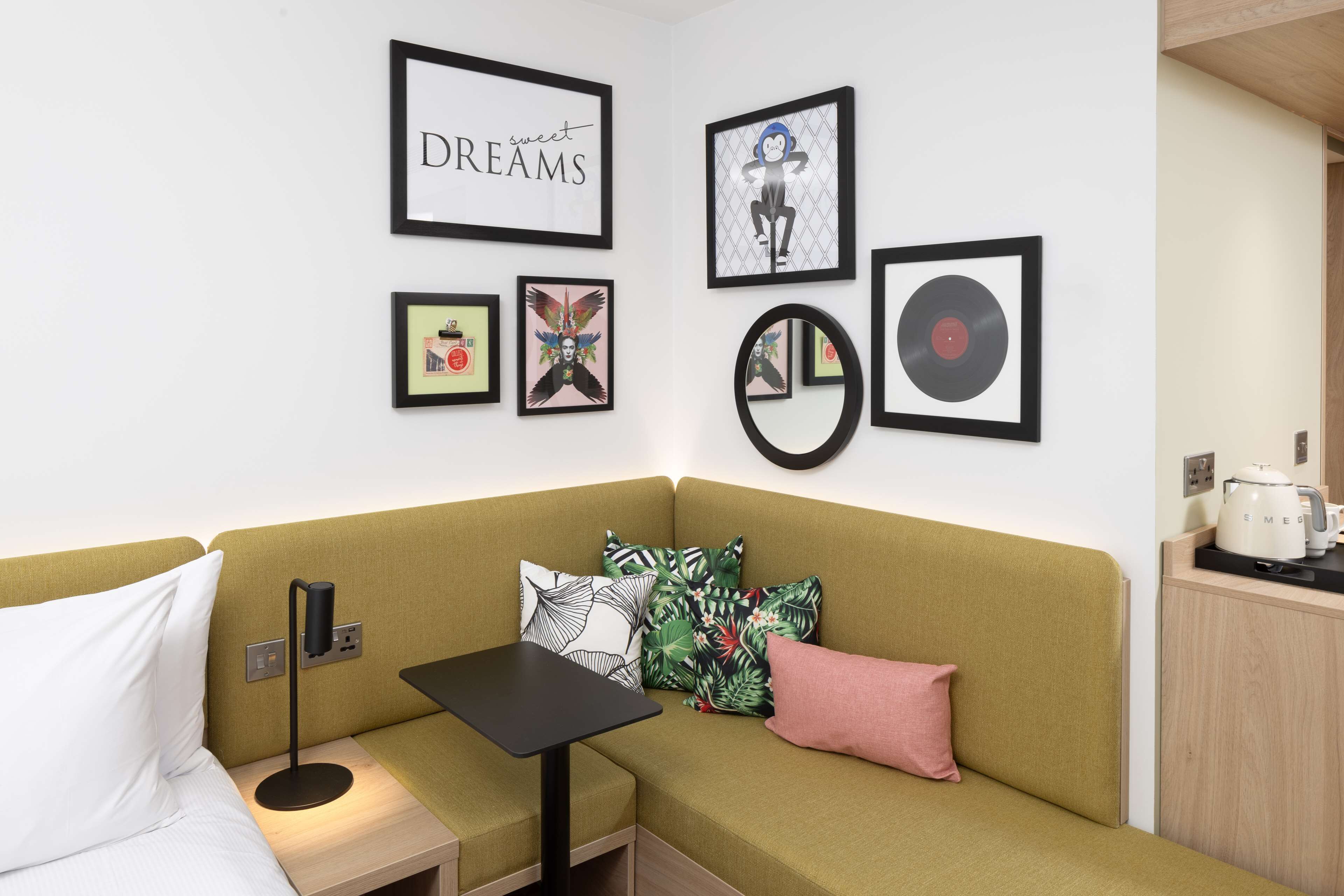 Een hoekappartement met een groene bank, een kleine tafel, een wandspiegel en kunstwerken, waaronder een vinylplaat en een bord met de tekst 'sweet DREAMS'.
