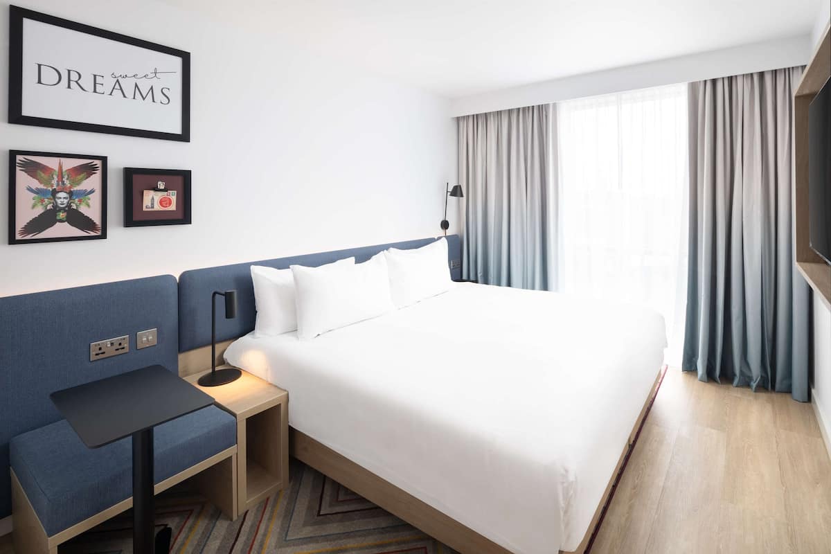 A hotel room with a large bed, a small desk, a nightstand, a wall-mounted TV, and a window with curtains.