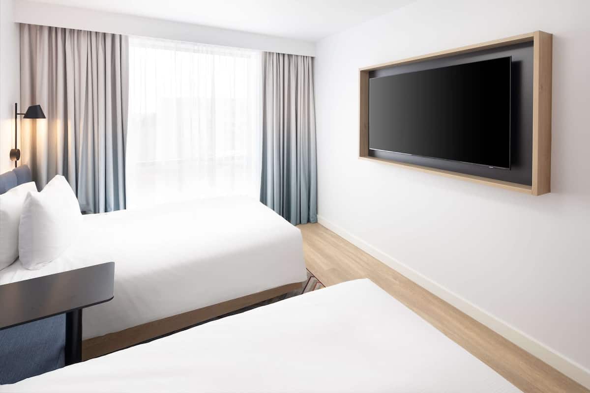 A hotel room with a bed, a desk, a TV mounted on the wall, and a window with curtains.
