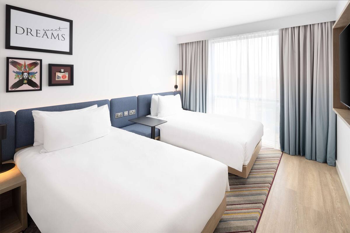 A hotel room with two beds, a desk, a TV, and a large window with curtains.
