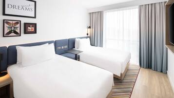 A hotel room with two beds, a desk, a TV, and a large window with curtains.