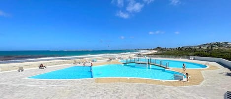 A pool area with multiple swimming pools, a beach, and a clear blue sky.