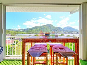 A balcony with two cushioned chairs, a table with a basket and a pot, and a view of a lake and mountains. - Luxury Studio in Mont Vernon Near Orient Bay Beach (Saint-Martin)