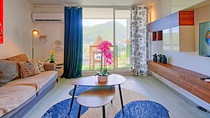Un salon moderne avec un canapé, une table ronde agrémentée d’une plante en pot, un téléviseur fixé au mur et un balcon offrant une vue sur la verdure.