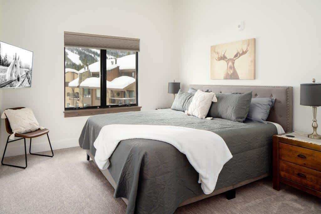 A bedroom with a large bed, a window with a view of buildings, a chair, and a nightstand.