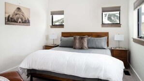 A bedroom with a large bed, two bedside tables, a headboard, and two windows with blinds.