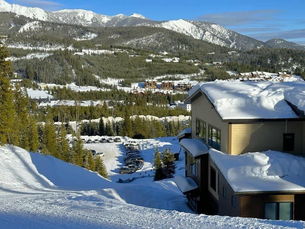 A modern building with a snow-covered roof, surrounded by a mountainous landscape and a parking area with several cars.