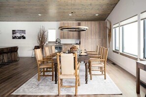 Uma área de refeições com mesa e cadeiras de madeira, luminária pendente e vista para a cozinha e janelas.