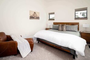 Um quarto com cama, uma poltrona de couro marrom, manta branca e duas mesas de cabeceira com abajures.
