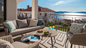 Un balcón con un sofá, sillas de mimbre, una mesa con una revista y uvas, y vista al mar.