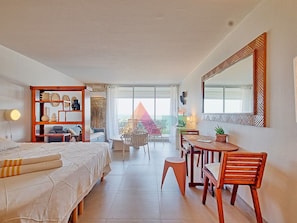 Habitación de hotel con una cama, un escritorio, una silla, una estantería y un ventanal grande.