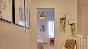 Een gang met een witte stoel, een hangplant en kunstwerken in lijsten aan de muur.