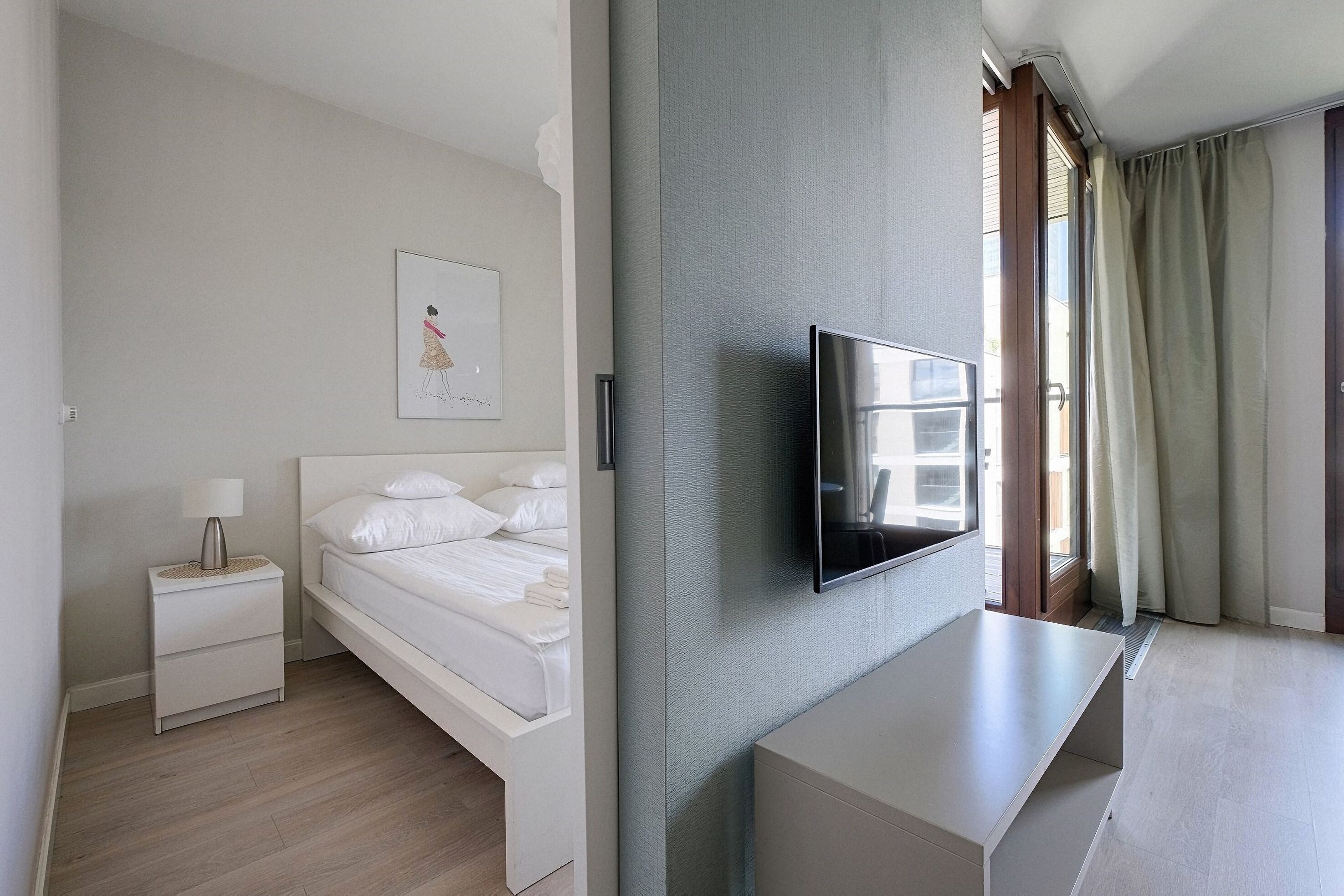 A modern hotel room with a bed, a TV mounted on the wall, a bedside table, and a window with curtains.