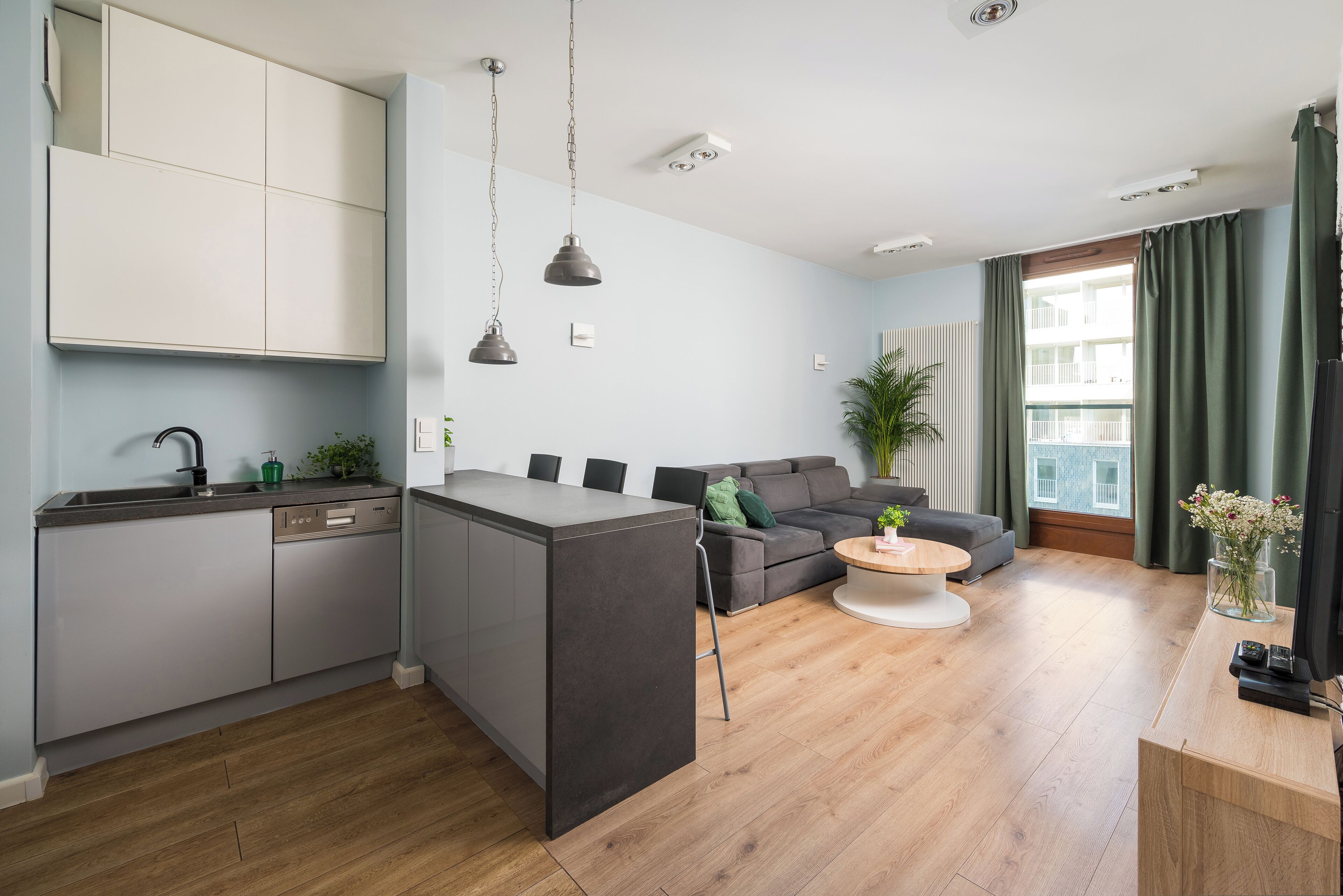 Eine moderne Wohnung mit Küche, Essbereich und Wohnzimmer. Sie verfügt über eine zentrale Kücheninsel, eine Sofagarnitur und ein großes Fenster mit Vorhängen.