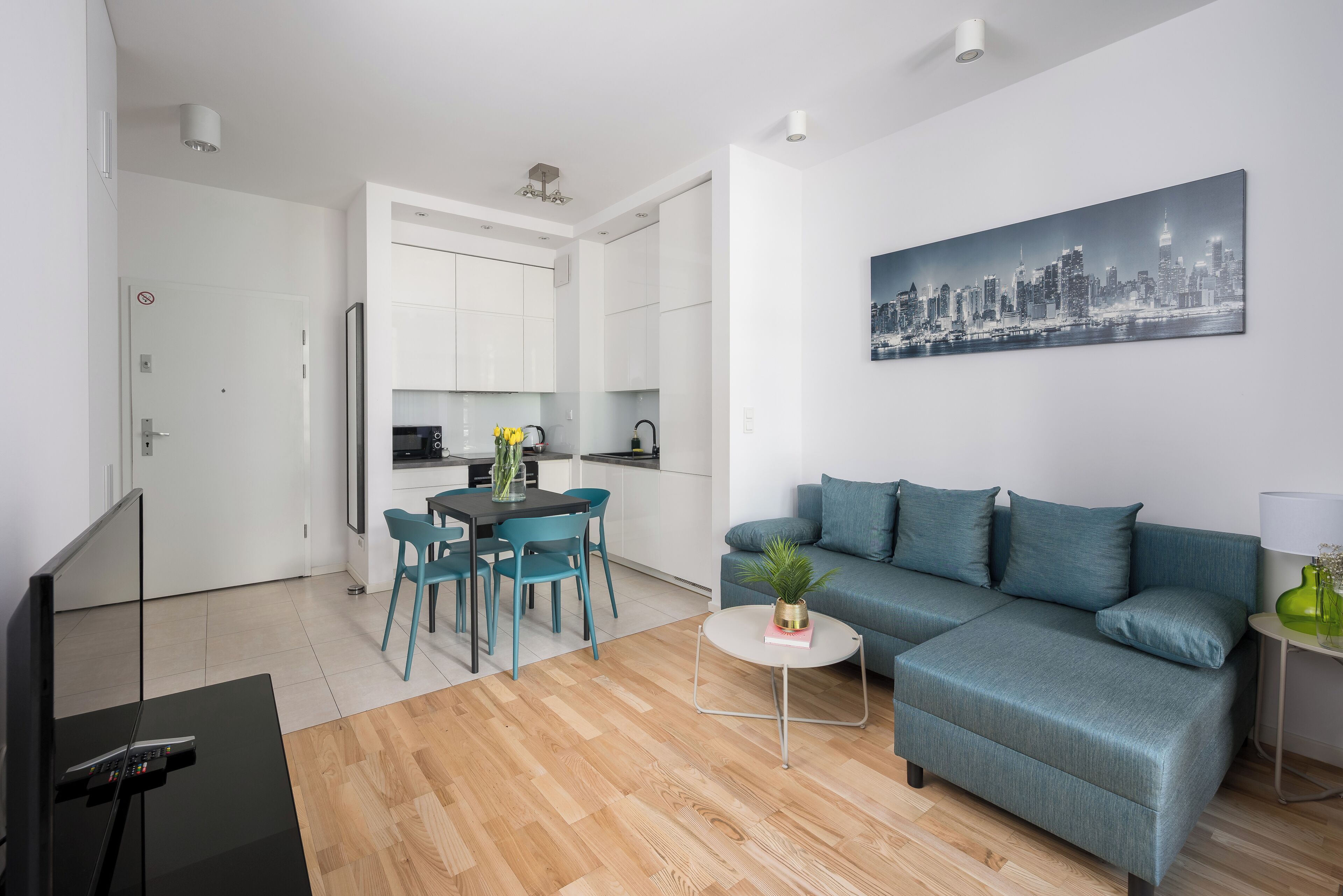 Una sala moderna con un sofá, una mesa de comedor y sillas. Hay una zona de cocina con armarios y un refrigerador. En la pared, un cuadro con una vista urbana.