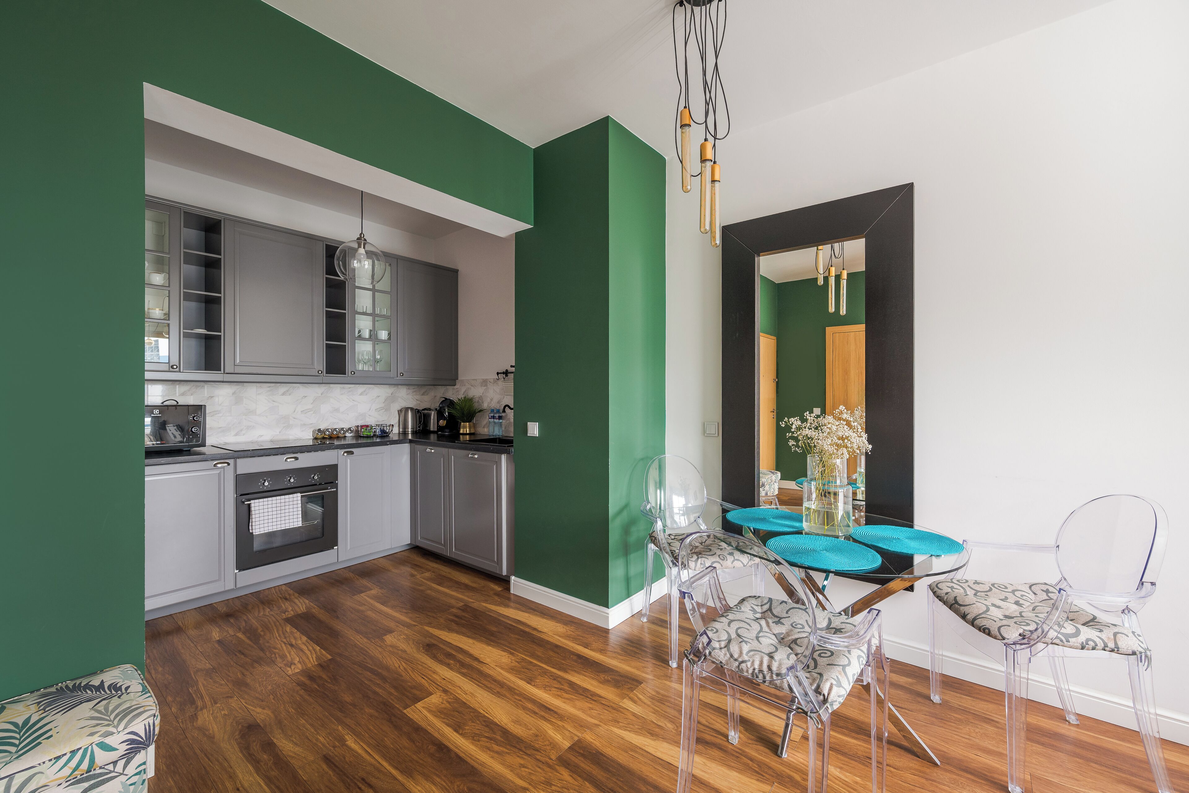 A modern kitchen with grey cabinets, a built-in oven, and a dining area with a clear glass table and chairs.